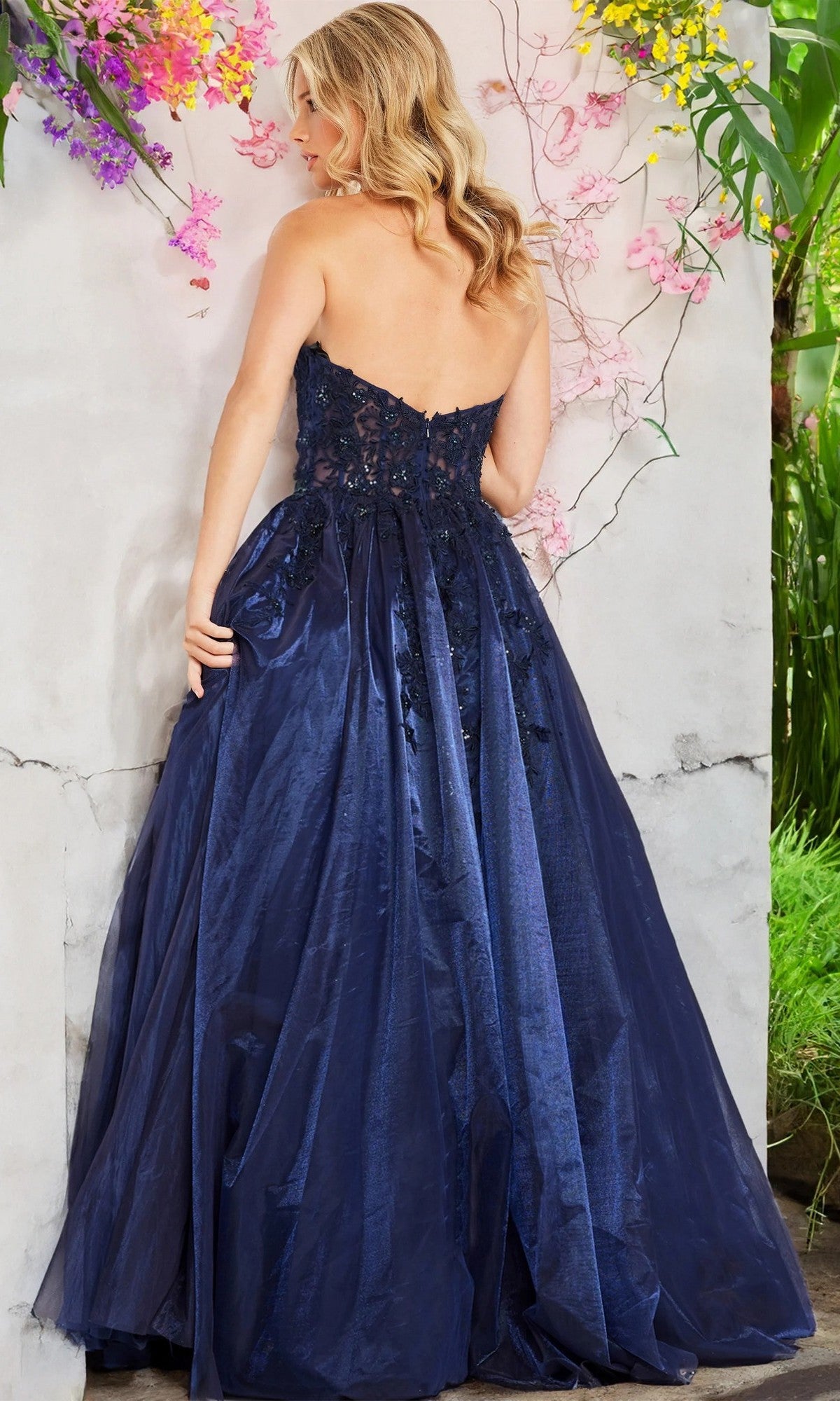 A woman stands with her back to the camera, wearing the Long Prom Dress: Jovani 00915—an elegant strapless navy blue gown with a tulle ballgown skirt and a delicately lace-embroidered bodice—beside a wall adorned with pink and yellow flowers.